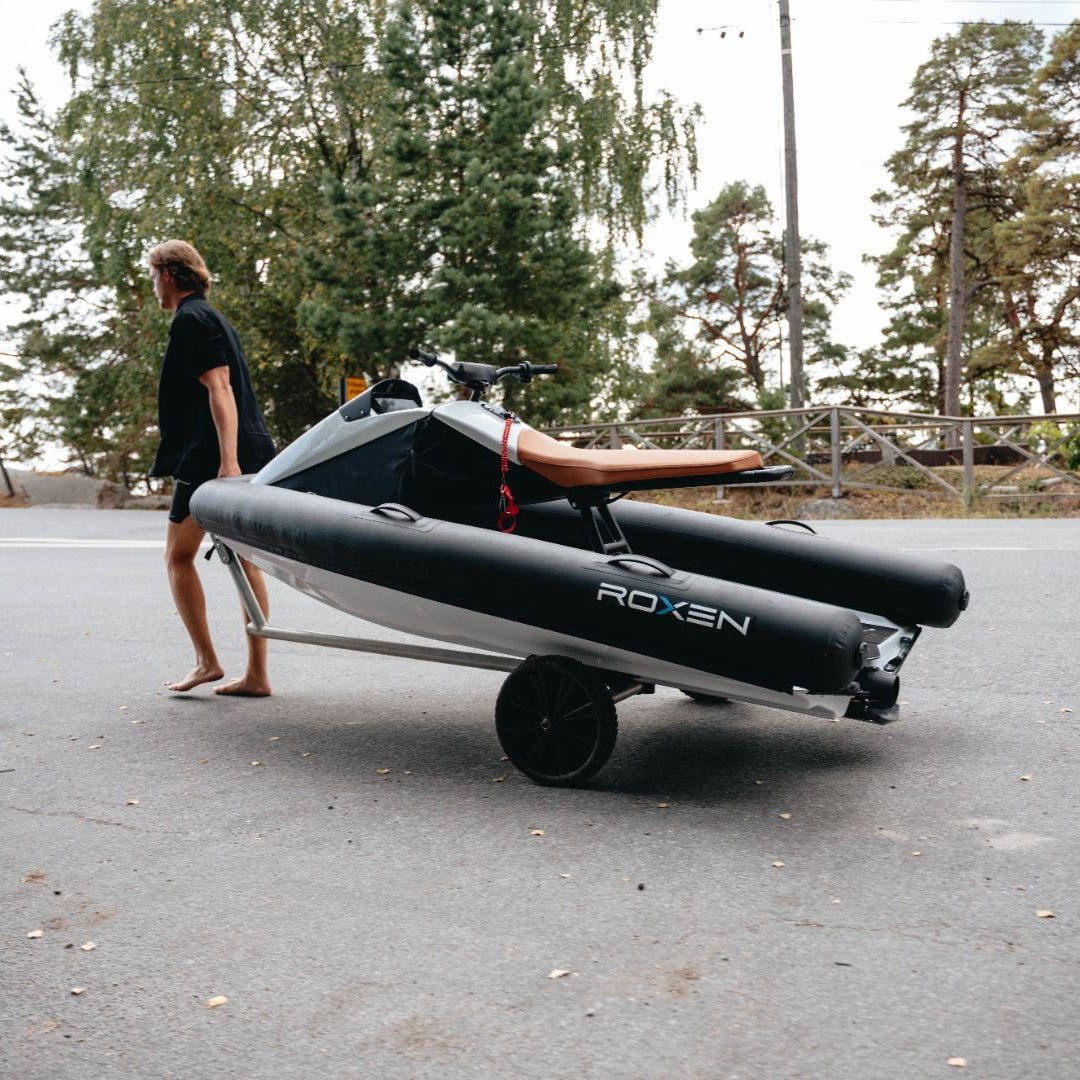 A man pulling a ROXEN electric watercraft on a small wheeled trailer along a tree-lined road